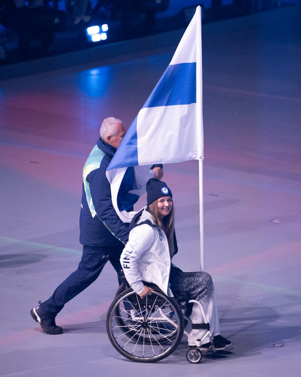 Nette Kiviranta kuljettamassa pyörätuoliinsa kiinnitettyä Suomen lippua läpi lavan.